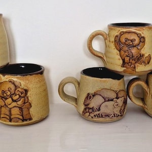 May include: A collection of eight tan ceramic mugs with dark brown rims and handles. Each mug features a unique, raised relief design of various animals, including dogs, bears, hippos, mice, and rabbits, rendered in a monochromatic brown tone.
