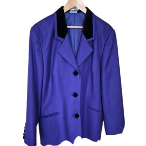 May include: A purple blazer with a black velvet collar and black buttons. The blazer has two front pockets and is hanging on a wooden hanger. The sleeves have black buttons at the cuffs.