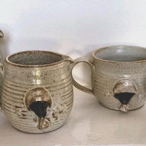 May include: A set of four ceramic vessels including one pitcher and three mugs, all featuring a speckled beige and brown glaze with horizontal ribbed textures and a distinct raised circular emblem with a black center on the front of each piece.