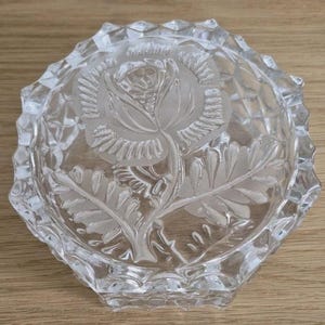 May include: Clear glass ashtray with a detailed rose design etched in the center. The ashtray has a faceted, octagonal shape with a textured rim. The rose is depicted with intricate leaves and petals.
