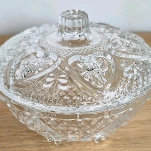 May include: A clear, vintage glass candy dish with a lid. The dish features a heart-shaped design and diamond-cut detailing. The lid has a textured knob for easy handling. The dish is sitting on a wooden surface.