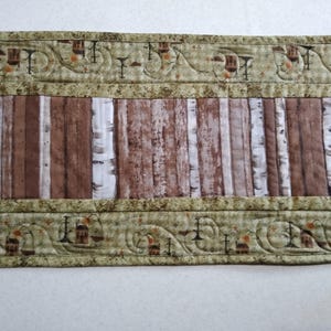 May include: A quilted table runner featuring a rustic wood plank design in shades of brown and white. The edges are bordered with a decorative pattern in shades of green and brown, adding a touch of elegance to the country-style decor.
