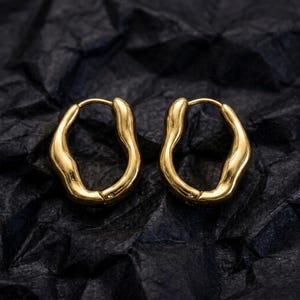 May include: A pair of gold-toned hoop earrings featuring an irregular, organic, and wavy silhouette. The earrings have a polished, reflective metallic surface and a continuous, closed-loop design with a smooth, undulating texture throughout the metal.