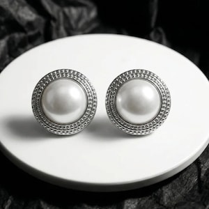 May include: A pair of round stud earrings featuring a central white pearl surrounded by a silver-toned metal border with a textured, beaded edge. The earrings are circular in shape and display a polished, reflective finish on the pearl surface.