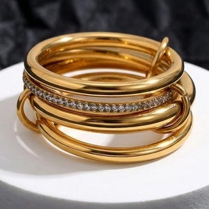 May include: A gold-toned multi-band ring featuring several smooth, polished circular bands stacked together. One of the central bands is embedded with a row of small, clear, sparkling stones that encircle the entire circumference of the ring.