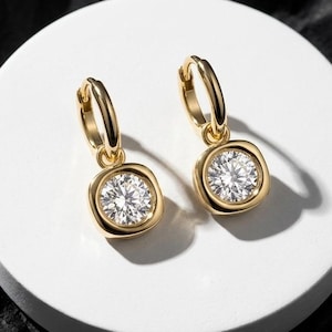 May include: A pair of gold hoop earrings featuring a dangling square-shaped charm with rounded corners. Each charm is bezel-set with a single round brilliant-cut clear gemstone that reflects light, creating a bright and polished appearance.