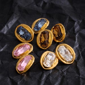 May include: A collection of seven oval-shaped earrings featuring gold-toned metal frames surrounding rectangular faceted crystals in various colours, including blue, brown, pink, and clear, arranged in a cluster on a dark textured surface.