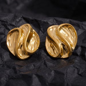 May include: A pair of gold-toned earrings featuring a rounded, abstract shape with a polished, reflective surface and deep, undulating grooves that create a sense of movement and texture across the metal form of each individual earring piece.