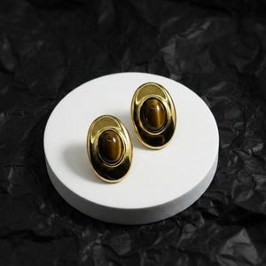 May include: A pair of oval-shaped earrings featuring a polished gold-tone metal frame surrounding a smooth, cabochon-cut brown tiger's eye gemstone. The earrings have a classic, symmetrical design with a reflective finish on the metal surface.