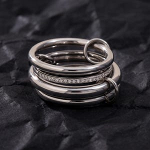 May include: A multi-band ring constructed from several polished silver-toned metal loops of varying thicknesses. One central band is embedded with a row of small, clear, sparkling stones, while the other smooth, rounded bands interlock around it.