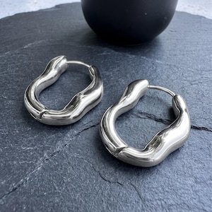 Op de afbeelding: Een paar zilverkleurige oorringen met een onregelmatige, organische en golvende vorm. De oorbellen hebben een gepolijst, reflecterend metalen oppervlak en een scharniersluiting, met een glad, vloeiend ontwerp en afgeronde randen.