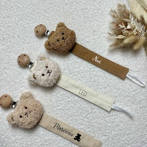 Peut inclure: Trois attaches-tétines pour bébés avec des têtes d'ours en peluche. Les attaches sont marron, crème et beige, avec les noms "Noé", "Léo" et "Marceau" imprimés sur les sangles. Chaque attache a une perle en bois et une boucle élastique blanche.