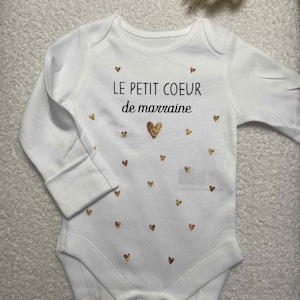 Peut inclure: Body blanc pour bébé avec des manches longues, orné de cœurs dorés scintillants et du texte français "LE PETIT COEUR de marraine". Le body a une fermeture à boutons-pression et est conçu pour les nourrissons.
