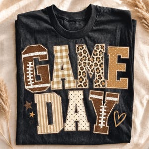 May include: A black short-sleeve t-shirt featuring the text GAME DAY in large block letters. The letters have varied patterns including brown football textures, beige gingham, leopard print, polka dots, and stripes, with small stars and a heart.