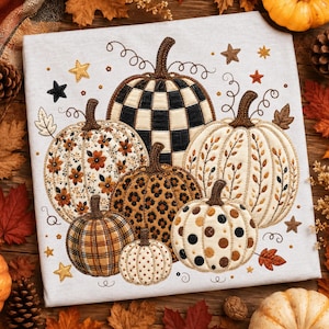 May include: A light gray t-shirt featuring an embroidered design of six pumpkins with various patterns including black and white checkered, floral, leopard print, plaid, and polka dot, accented with small embroidered stars and swirling vines.