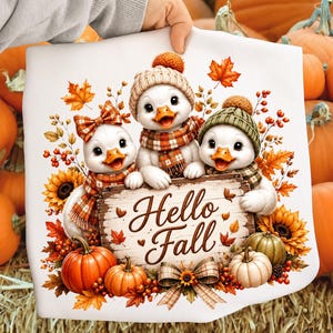 May include: A white t-shirt featuring a graphic of three cartoon ducks wearing knit hats and plaid scarves, holding a wooden sign that reads Hello Fall. The design is surrounded by orange pumpkins, sunflowers, red berries, and autumn leaves.