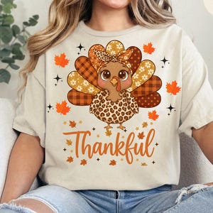 Könnte beinhalten: Beiges T-Shirt mit einem Cartoon-Truthahn-Design. Der Truthahn hat ein gemustertes Kopftuch und einen Leopardenmuster-Oberteil. Das Wort "Thankful" steht in orangefarbener Schrift darunter, mit Herbstblättern und Sternen.
