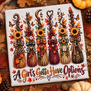 May include: A white t-shirt featuring a graphic of six decorative broomsticks adorned with autumn leaves, sunflowers, and various bows. The text "A Girl's Gotta Have Options" is printed in a colorful script font at the bottom of the design.