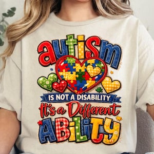 Puede incluir: Camiseta color crema con un diseño gráfico colorido. El diseño presenta la palabra "Autismo" en letras azules y rojas, un rompecabezas en forma de corazón y la frase "No es una discapacidad, es una capacidad diferente."