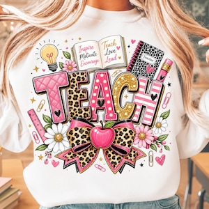 May include: A white sweatshirt featuring a graphic print of the word TEACH in pink quilted, leopard print, and gold glitter textures. The design includes an open book with text, a leopard print notebook, a pencil, an apple, and flowers.