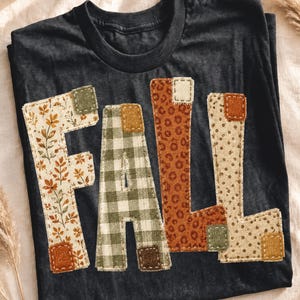 Op de afbeelding: Een zwart T-shirt met ronde hals en het woord FALL in grote, opgestikte stoffen letters. Elke letter is gemaakt van verschillende stoffen met patronen, waaronder herfstbladeren, groen-witte vichy-ruitjes, luipaardprint en beige stippen.
