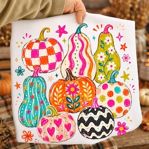May include: A white t-shirt featuring a colorful graphic print of various pumpkins. The pumpkins display diverse patterns including pink and orange checkers, wavy stripes, floral motifs, polka dots, and hearts, accented by small stars and flowers.