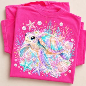 Könnte beinhalten: Ein leuchtend pinkfarbenes T-Shirt mit kurzen Ärmeln und einer bunten Grafik einer Meeresschildkröte, umgeben von Seesternen, Muscheln und Blasen. Die Schildkröte hat einen schillernden, mehrfarbigen Panzer und große Augen vor einer Unterwasserkulisse.
