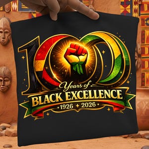 May include: Black pillow with a gold-outlined design celebrating "100 Years of Black Excellence." The design includes a raised fist, the years 1926-2026, and red, yellow, and green accents. The pillow is held by a hand.