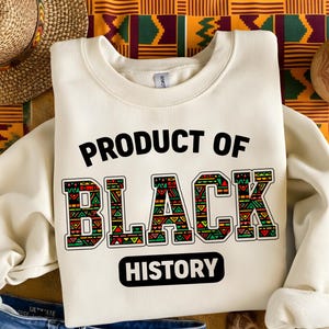May include: Cream-colored sweatshirt with the text "PRODUCT OF BLACK HISTORY" in black and colorful letters. The letters are decorated with geometric patterns in red, green, and yellow. The sweatshirt is folded on a patterned fabric.