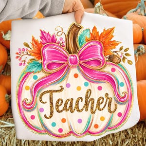 May include: A white t-shirt featuring a graphic of a cream-colored pumpkin decorated with a large bright pink bow, colorful polka dots, and autumn leaves. The word Teacher is written across the center in gold glitter script lettering.