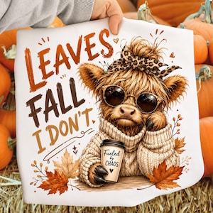 Può includere: Una t-shirt bianca con la grafica di una mucca Highland pelosa con occhiali da sole e una fascia leopardata. La mucca indossa un maglione beige e tiene in mano un bicchiere di caffè con scritto 'Fueled by CHAOS'. Testo: 'LEAVES FALL I DON'T'.