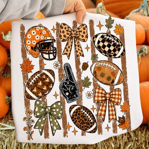 May include: A white t-shirt featuring a graphic print of football-themed elements including an orange daisy-patterned helmet, three bows with leopard, plaid, and polka dot patterns, three footballs with various textures, and a foam finger.