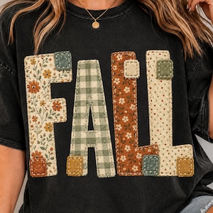 Op de afbeelding: Een zwart T-shirt met korte mouwen en het woord FALL in grote blokletters. Elke letter is gevuld met een ander stofpatroon, waaronder bloemenprints in oranje en crème, een groen-witte ruitjesstof en stippen.