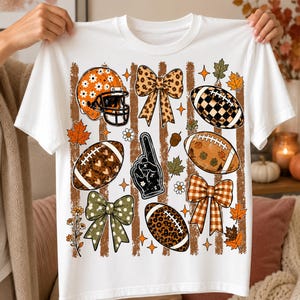 May include: A white t-shirt featuring a graphic print of football-themed elements including a helmet with daisies, a foam finger, various bows, and footballs with leopard, plaid, and heart patterns, all set against vertical brown stripes and autumn leaves.