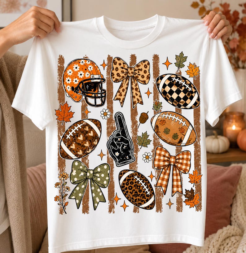 May include: A white t-shirt featuring a graphic print of football-themed elements including a helmet with daisies, a foam finger, various bows, and footballs with leopard, plaid, and heart patterns, all set against vertical brown stripes and autumn leaves.