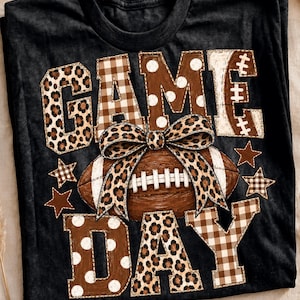 Könnte beinhalten: Ein schwarzes T-Shirt mit dem Schriftzug GAME DAY in großen Blockbuchstaben mit Leopardenmuster, braun-weißem Vichy-Karo und weißen Punkten. Ein brauner Football mit einer Leoparden-Schleife liegt mittig, umgeben von braunen Sternen.