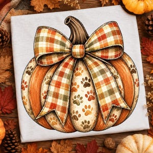Puede incluir: Una camiseta gris claro con una ilustración gráfica de una calabaza naranja decorada con un gran lazo a cuadros en tonos naranja, verde y blanco. La superficie de la calabaza está cubierta con un patrón repetitivo de pequeñas huellas de patas.