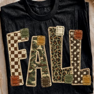 Op de afbeelding: Een zwart t-shirt met korte mouwen en het woord FALL in grote blokletters. Elke letter heeft een uniek patroon: een bruin-wit schaakbordpatroon, groen camouflage, luipaardprint en witte stippen op een bruine achtergrond.
