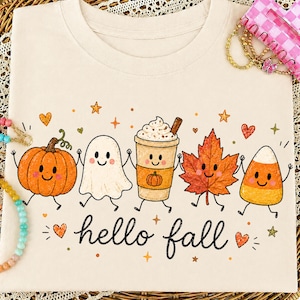 May include: A cream-colored t-shirt featuring a graphic print of a smiling pumpkin, a ghost, a coffee cup, a maple leaf, and a piece of candy corn holding hands. The text "hello fall" is printed in black script below the characters with small stars and hearts.