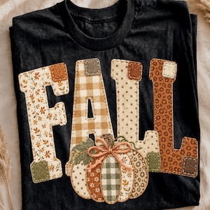 May include: A black crew neck t-shirt featuring the word FALL in large, fabric-appliqued letters. Each letter is made of different patterned fabrics in shades of brown, tan, and cream. A pumpkin graphic sits at the base with a twine bow.