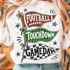 May include: A white sweatshirt featuring a graphic of three stacked pennant flags on a pole. The top brown flag says FOOTBALL, the middle green flag says TOUCHDOWN with yard lines, and the bottom checkered flag says GAMEDAY, accented by stars.