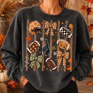 May include: A charcoal gray crewneck sweatshirt featuring a graphic print of football-themed elements including a helmet, footballs, bows, and a foam finger. The design incorporates autumn leaves, stars, and daisies over vertical brown stripes.