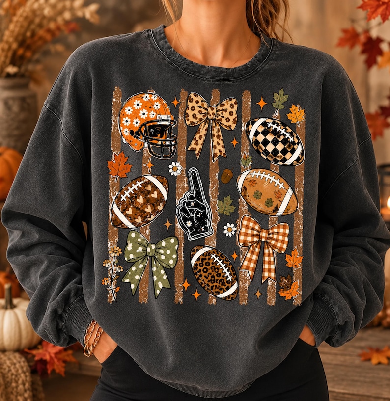 May include: A charcoal gray crewneck sweatshirt featuring a graphic print of football-themed elements including a helmet, footballs, bows, and a foam finger. The design incorporates autumn leaves, stars, and daisies over vertical brown stripes.
