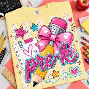 May include: A pale yellow t-shirt featuring a graphic print of a yellow pencil with a large pink bow, a piece of lined notebook paper, and the text "pre-k" in glittery pink script. The design includes pink and blue stars and hearts.