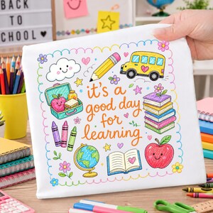 以下が含まれることがあります： 黄色いスクールバス、鉛筆、地球儀、本、リンゴなどの学用品のカラフルなイラストが描かれた白い布製の正方形。テキストには「it's a good day for learning」と書かれており、花のスカラップ模様の縁取りで囲まれています。
