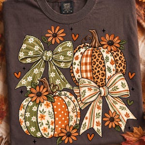 Può includere: Una t-shirt grigio scuro con una stampa grafica di due zucche decorate con motivi che includono maculato, quadri e pois. Il design include grandi fiocchi verdi e a righe, fiori arancioni e piccoli cuori arancioni.