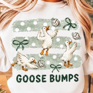 Peut inclure: Un sweat-shirt blanc orné d'un motif représentant trois oies blanches jouant au volley-ball. Le design comprend des coups de pinceau horizontaux vert sauge à pois blancs, des nœuds vert foncé et le texte GOOSE BUMPS en lettres capitales.