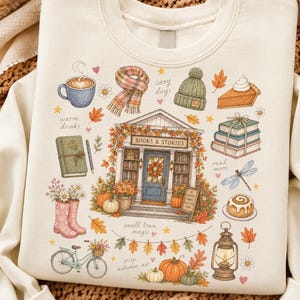 May include: A cream-colored sweatshirt featuring a colorful graphic print of a bookstore storefront labeled "BOOKS & STORIES" surrounded by autumn-themed illustrations including a coffee mug, plaid scarf, knit hat, pumpkin pie, books, and a lantern.