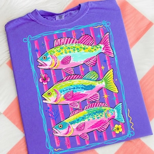 Può includere: Una maglietta viola con una grafica colorata di tre pesci. I pesci hanno dettagli rosa, gialli, verdi e blu. Il design è incorniciato da un bordo azzurro e strisce verticali. Sono inclusi anche elementi floreali e acquatici.