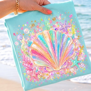 May include: A light blue beach towel featuring a colorful seashell design with a rainbow gradient. The seashell is surrounded by starfish, bubbles, and glitter. The towel is held by a person with a gold bracelet.